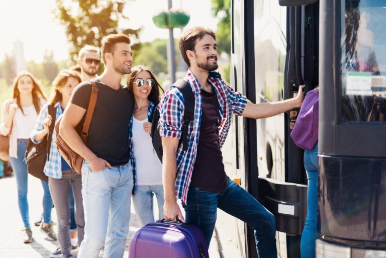 A group of tourists wearing bags are in line to get on a tour bus